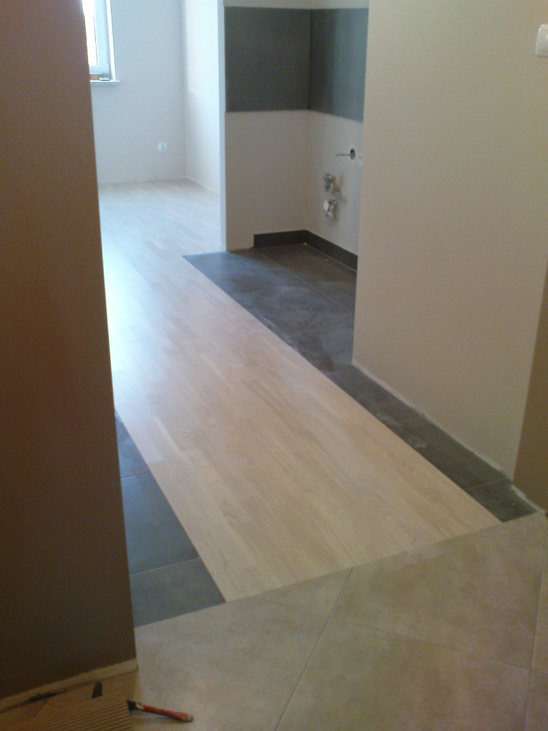 Fragment wnętrza z nową, jasną podłogą drewnianą przechodzącą w szare płytki, widoczne przygotowane miejsce na umywalkę, ściany w odcieniach bieli i beżu, częściowo pomalowane na szaro.